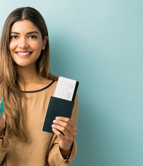 Happy young woman holding passport and boarding pass with globe while standing against blue background